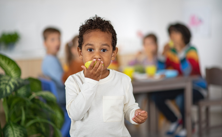 Multiracial small boy with group of children eating snack and looking at camera indoors at nursery school.