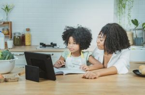 A single mother and African American daughter are teaching their children homework and studying online with their children by greeting friends and teachers or video calling with their father on his way home. By the way, the house prepares to cook food. in