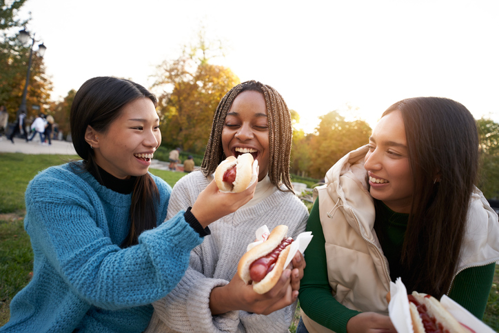 Group of happy smiling girls eating takeaway street food sitting on a bench in a nice city park.