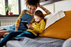 Quality childcare matters. African american woman baby sitter and caucasian cute little girl smiling, using tablet pc, sitting at home. Children education, leisure activities, babysitting concept