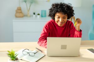 African american woman using laptop shopping online paying with gold credit card. Girl sitting at home buying on Internet enter credit card details. Online shopping ecommerce delivery service.