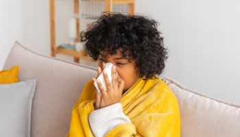 Unhappy sick young african american woman with cold sitting on sofa at home. Girl covering with blanket and blowing her nose in tissue. Medical and health concept. Cold flu allergy. Sick at home.