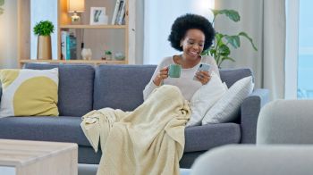 Coffee, phone and happy black woman on sofa in home living room, social media and reading news. Drink tea, smartphone and African person on couch in blanket, relax and typing email on internet app.