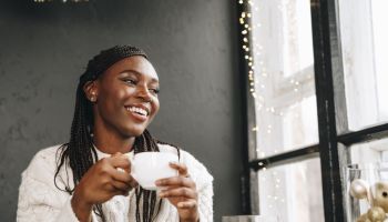 African woman in warm white sweater drinking hot coffee or tea at home