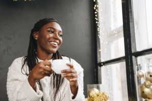 African woman in warm white sweater drinking hot coffee or tea at home