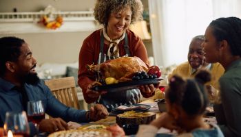 Happy black mature woman serving Thanksgiving turkey to her family at dining table.