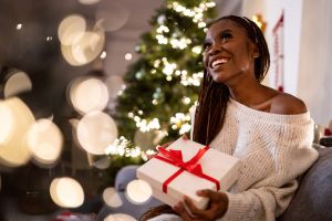 Young woman Black ethnicity holding an Christmas present while sitting on the sofa near the Christmas tree