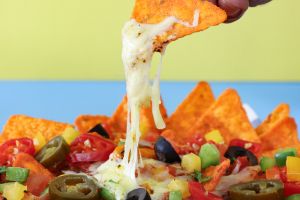 Full frame image of unrecognisable person holding nacho, plate of loaded nachos covered in tomato salsa, melted mozzarella cheese, tomatoes, black olives, bell peppers, Jalapeno peppers, red chillies, split yellow-blue background, focus on foreground