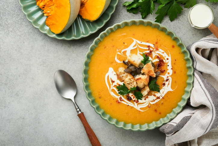 Pumpkin and carrot cream soup with herbs, seasonings and seeds in bowl on gray concrete table background in rustic style. Thanksgiving traditional autumn pumpkin cream soup. Top view.