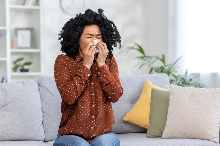 Sick young African-American woman sitting on sofa at home and wiping nose from runny nose with tissue, not feeling well