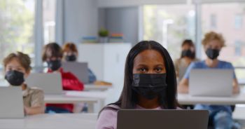 African-American schoolgirl put on medical mask using laptop during lesson looking at camera