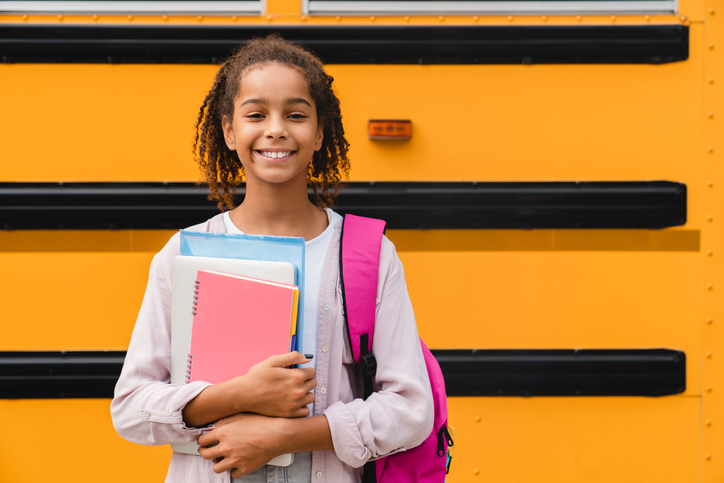 African-american girl teenager pupil student preparing to go to school after summer holidays holding books and notebooks standing next to the school bus.