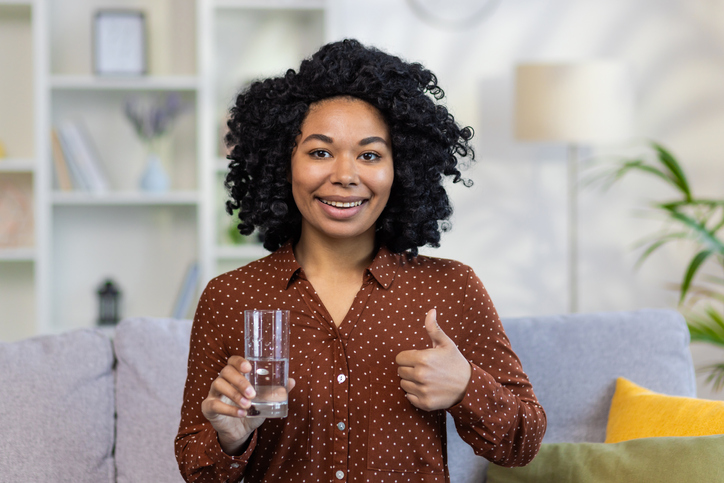 Portrait of an African-American woman sitting on the couch at home, holding a glass of water in her hands, pointing super finger, smiling and confidently looking at the camera