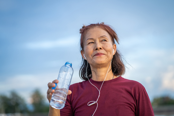 Healthy senior woman drinks water after exercise on a walkway by a pond in a park.