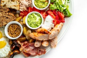 A vibrant breakfast platter featuring assorted breads, fresh vegetables, creamy avocado, tuna, cheeses, sausages, bacon, and eggs. Perfect for a hearty morning meal or brunch setting.