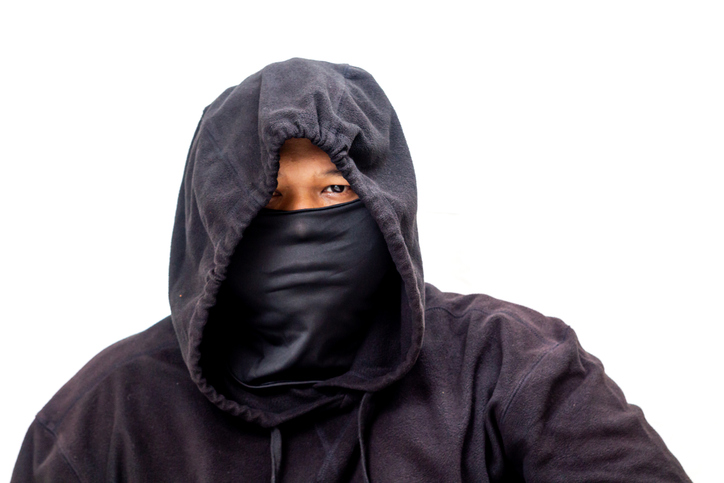 Mysterious individual in black hoodie and face mask stares intensely against clean white backdrop.