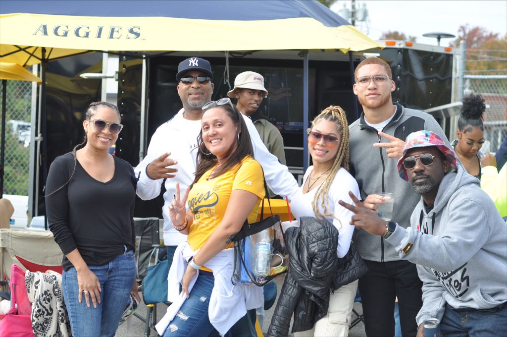 A group of people standing in front of a yellow Aggies umbrella, some wearing baseball caps and sunglasses, smiling and making hand gestures.