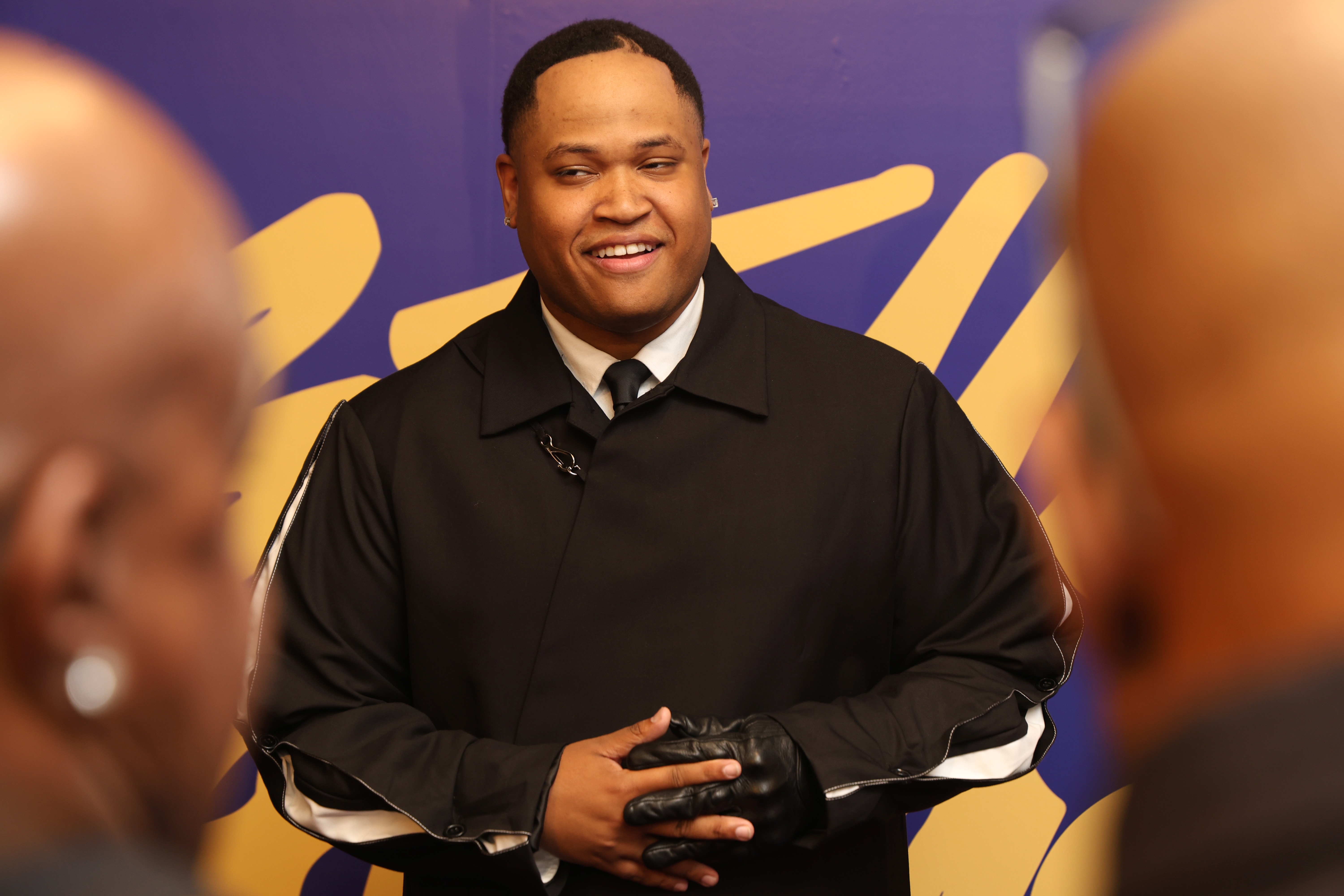 A smiling man in a black suit and tie standing in front of a purple background.