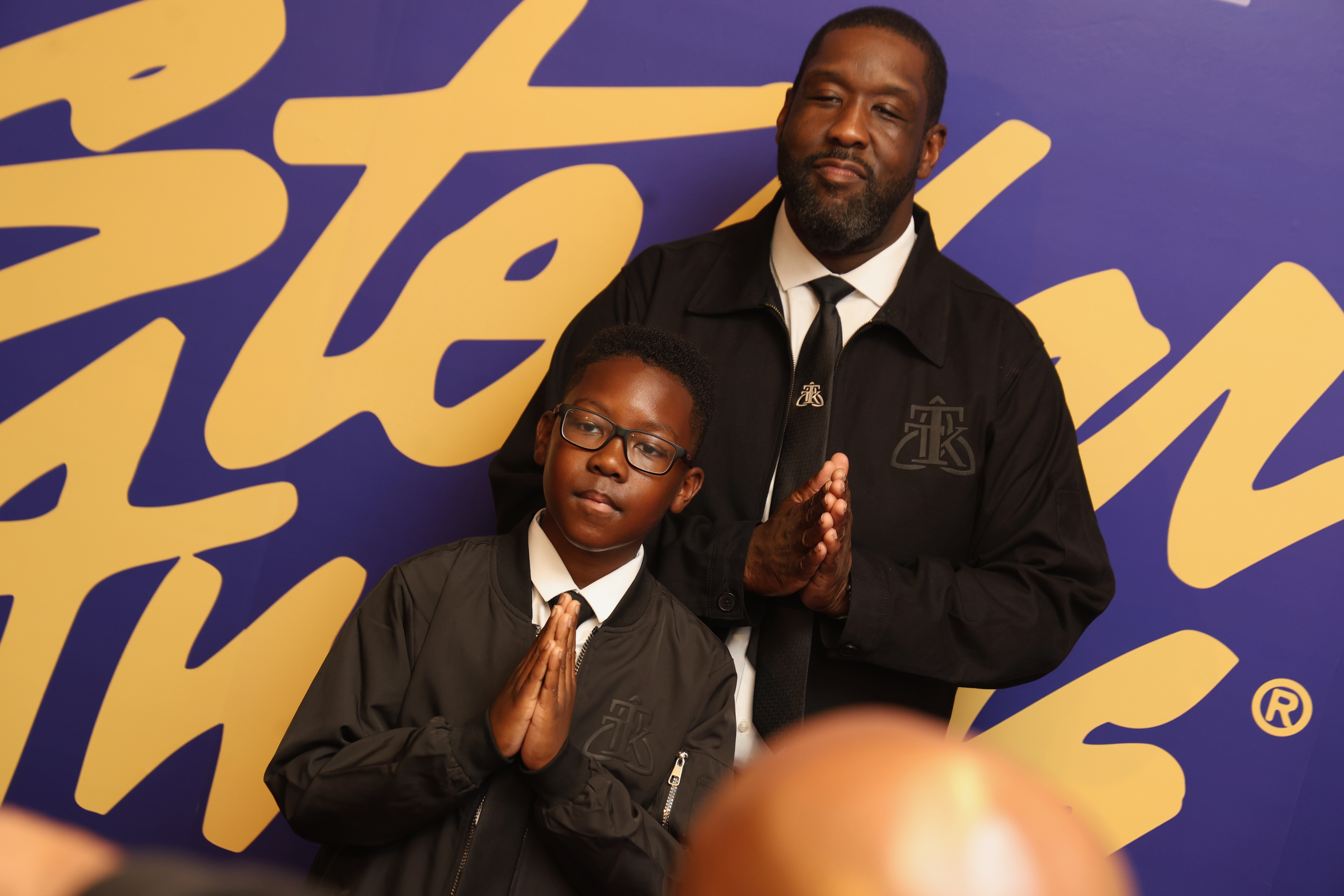 A man in a suit stands with a young boy in a school uniform, both with hands clasped in a gesture of prayer or reverence, in front of a colorful graphic background.