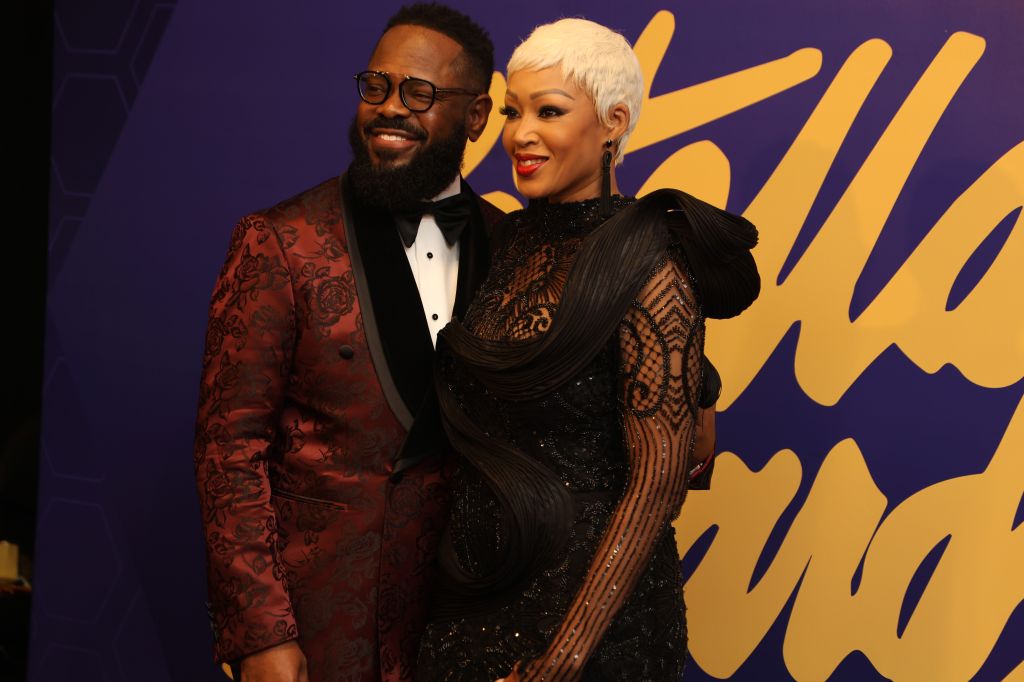 Two well-dressed individuals, a man in a red floral tuxedo jacket and a woman in a black sequined gown, posing together at an event.