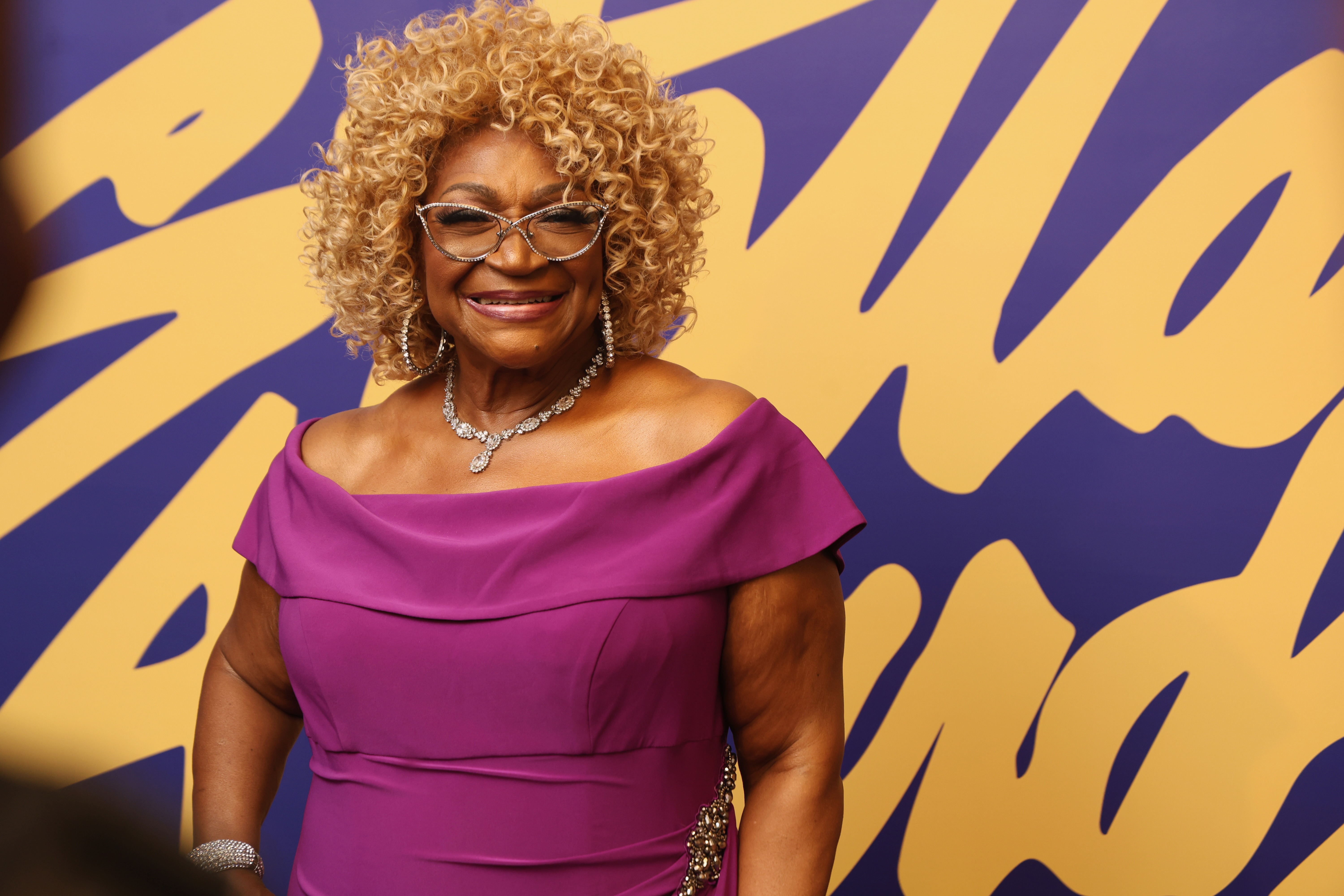 A smiling woman with curly blonde hair wearing a pink off-the-shoulder dress and large diamond jewelry stands in front of a purple and yellow abstract background.
