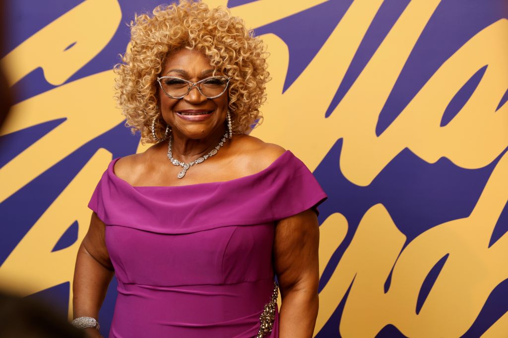 A smiling woman with curly blonde hair wearing a pink off-the-shoulder dress and large diamond jewelry stands in front of a purple and yellow abstract background.