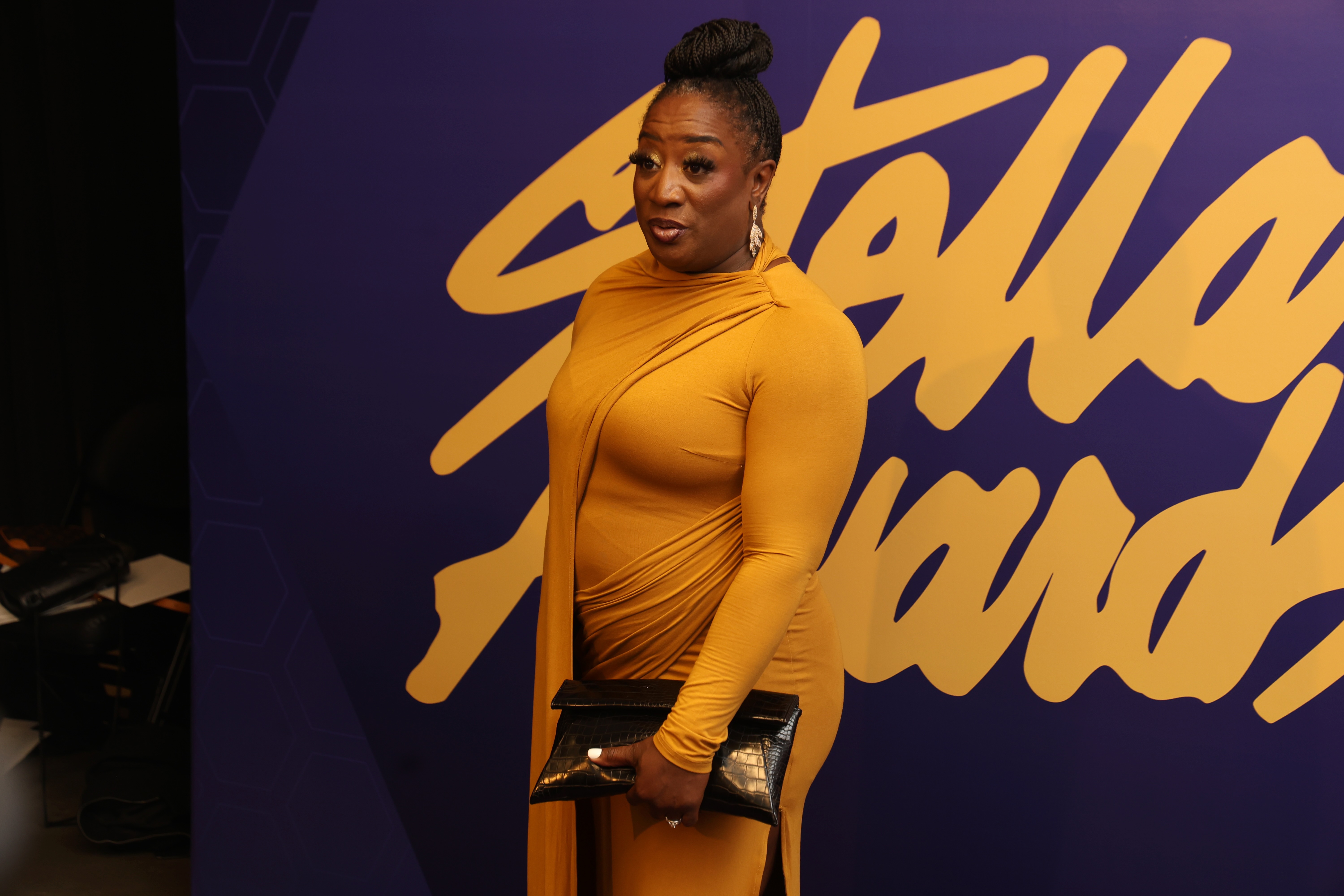 A person wearing a mustard-colored dress stands in front of a purple and yellow background.