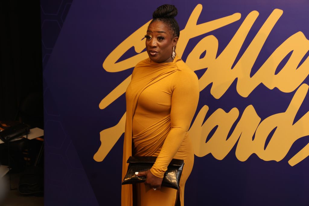 A person wearing a mustard-colored dress stands in front of a purple and yellow background.