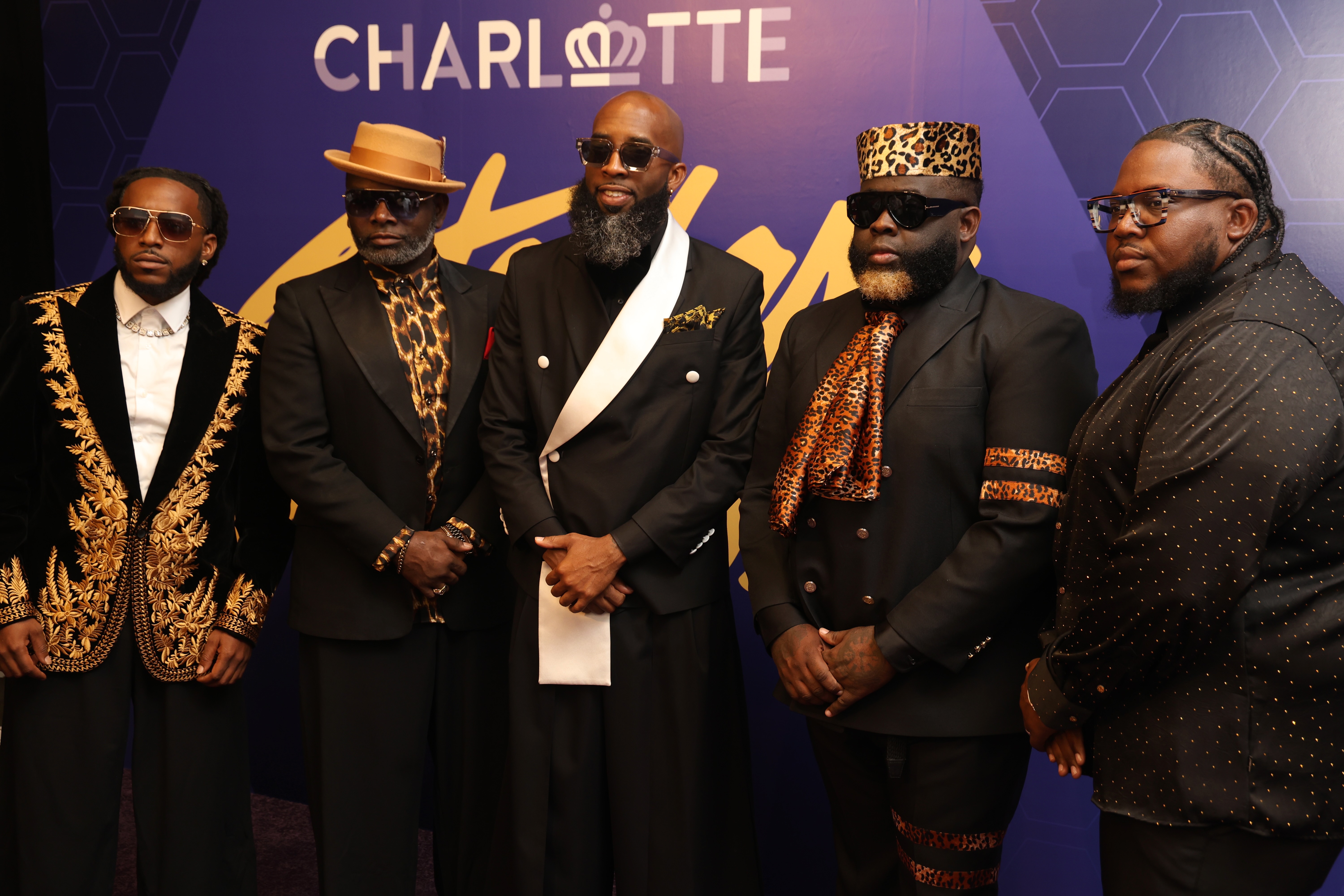Five men in formal attire, including leopard print and gold-accented suits, standing together in front of a "Charlotte" backdrop.