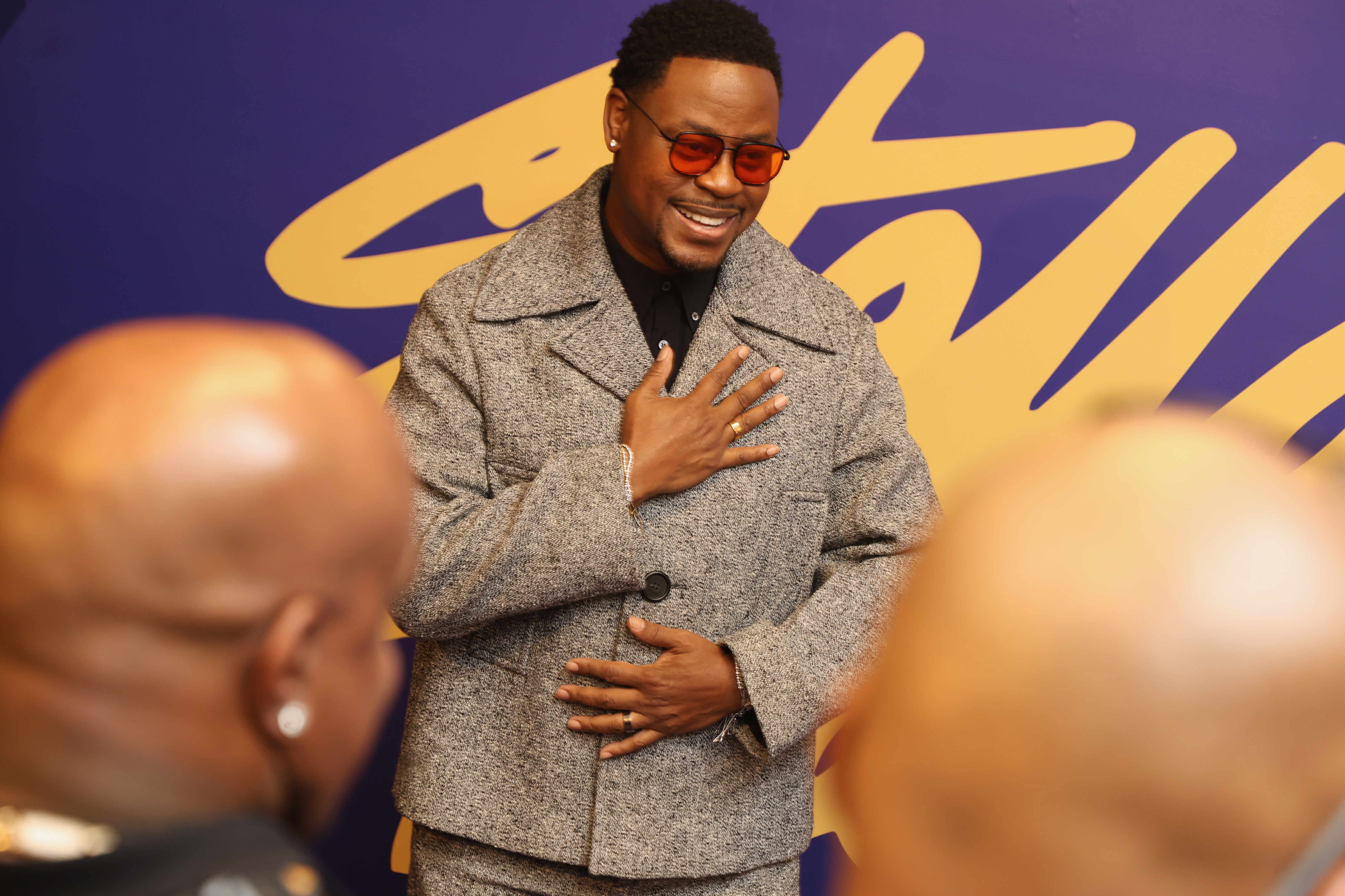 A smiling man wearing a gray coat and sunglasses stands in front of a yellow and purple background.