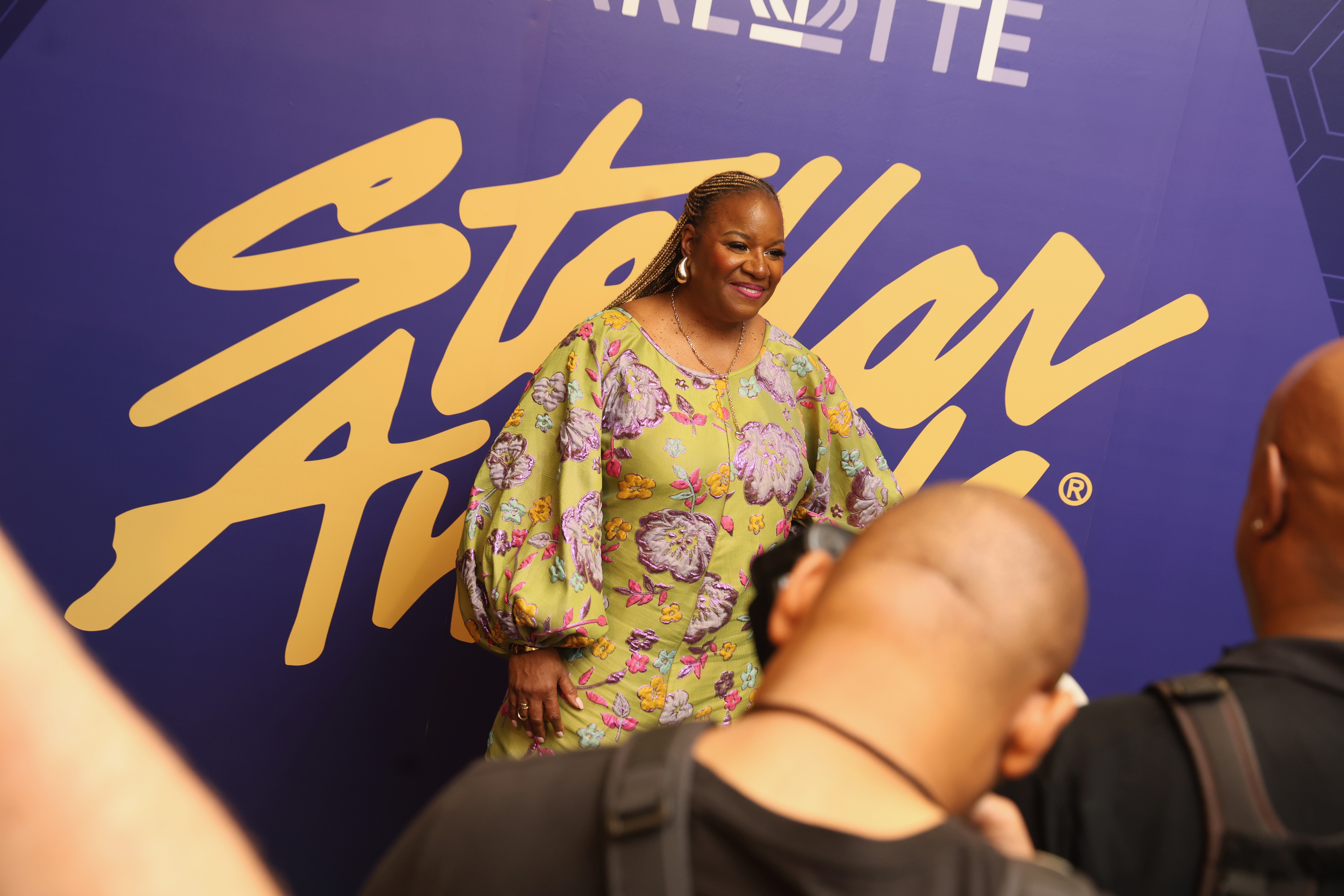 A woman wearing a bright floral dress stands in front of a large "Stella Artois" logo backdrop.