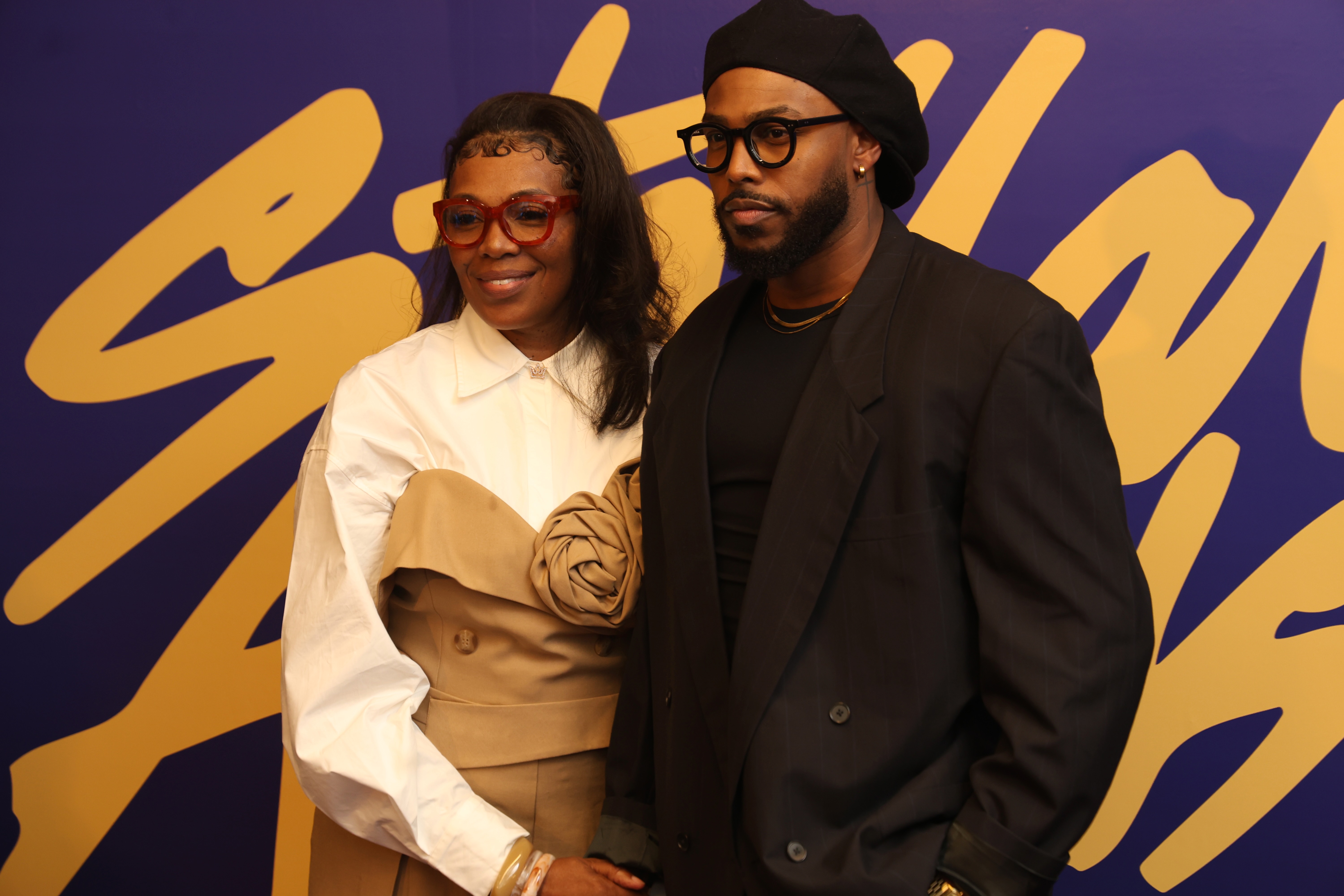 Two people wearing stylish eyeglasses posing in front of a vibrant purple and yellow background.