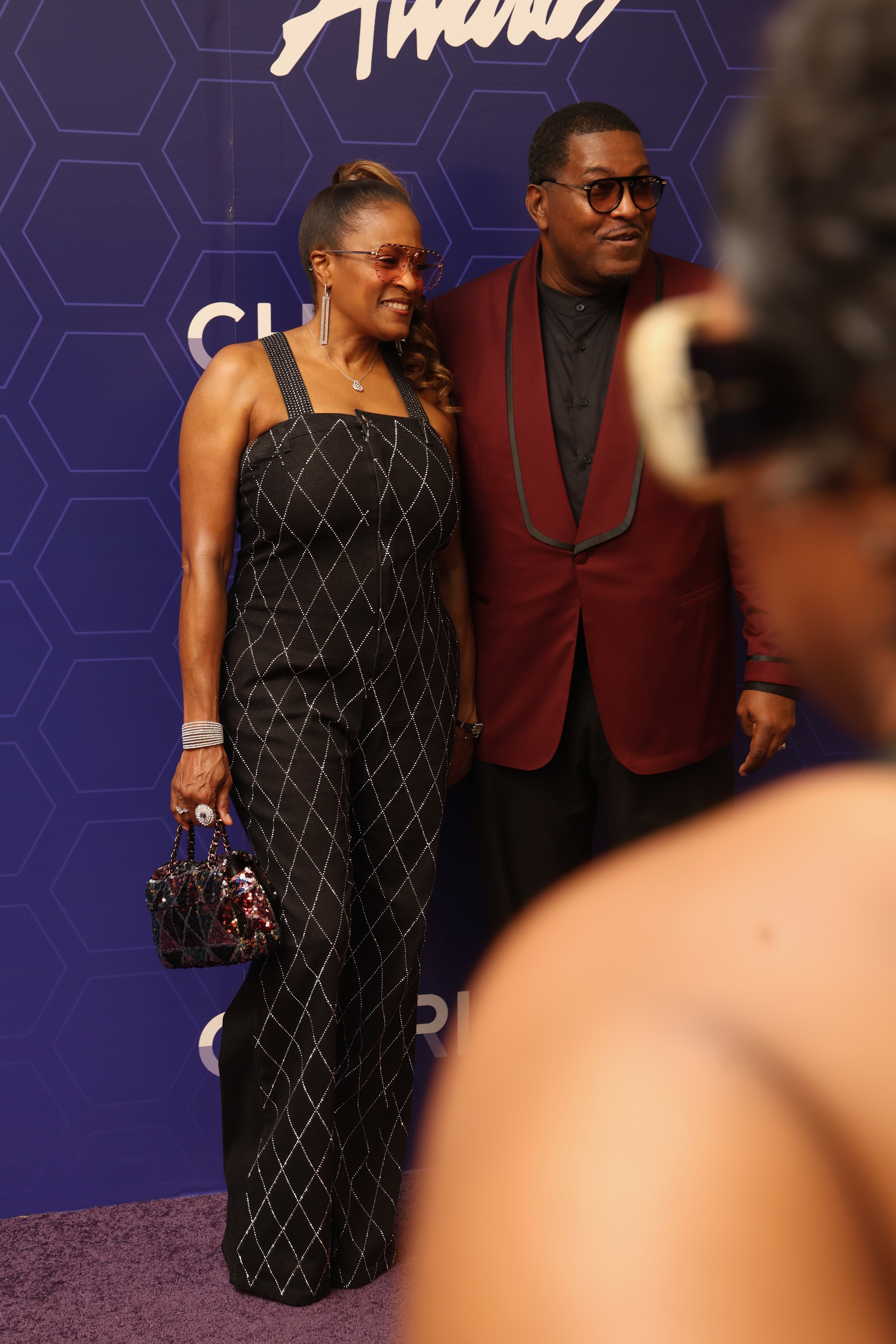 Two well-dressed individuals, a woman in a black and white patterned jumpsuit and a man in a red suit, posing together on a purple background with the text "Amani" visible.