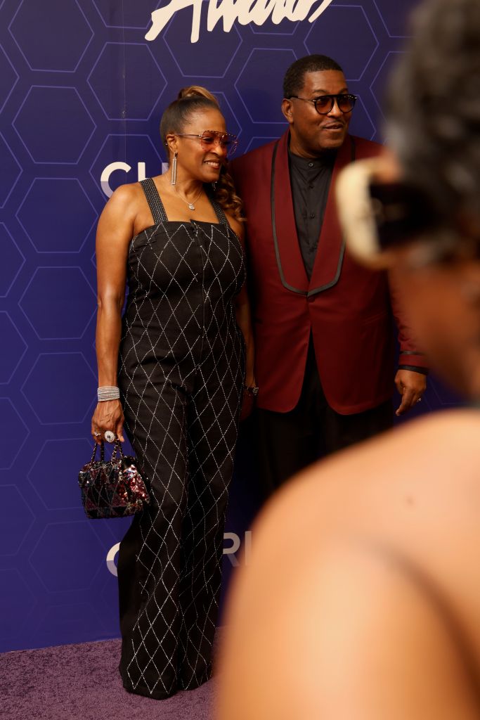 Two well-dressed individuals, a woman in a black and white patterned jumpsuit and a man in a red suit, posing together on a purple background with the text "Amani" visible.