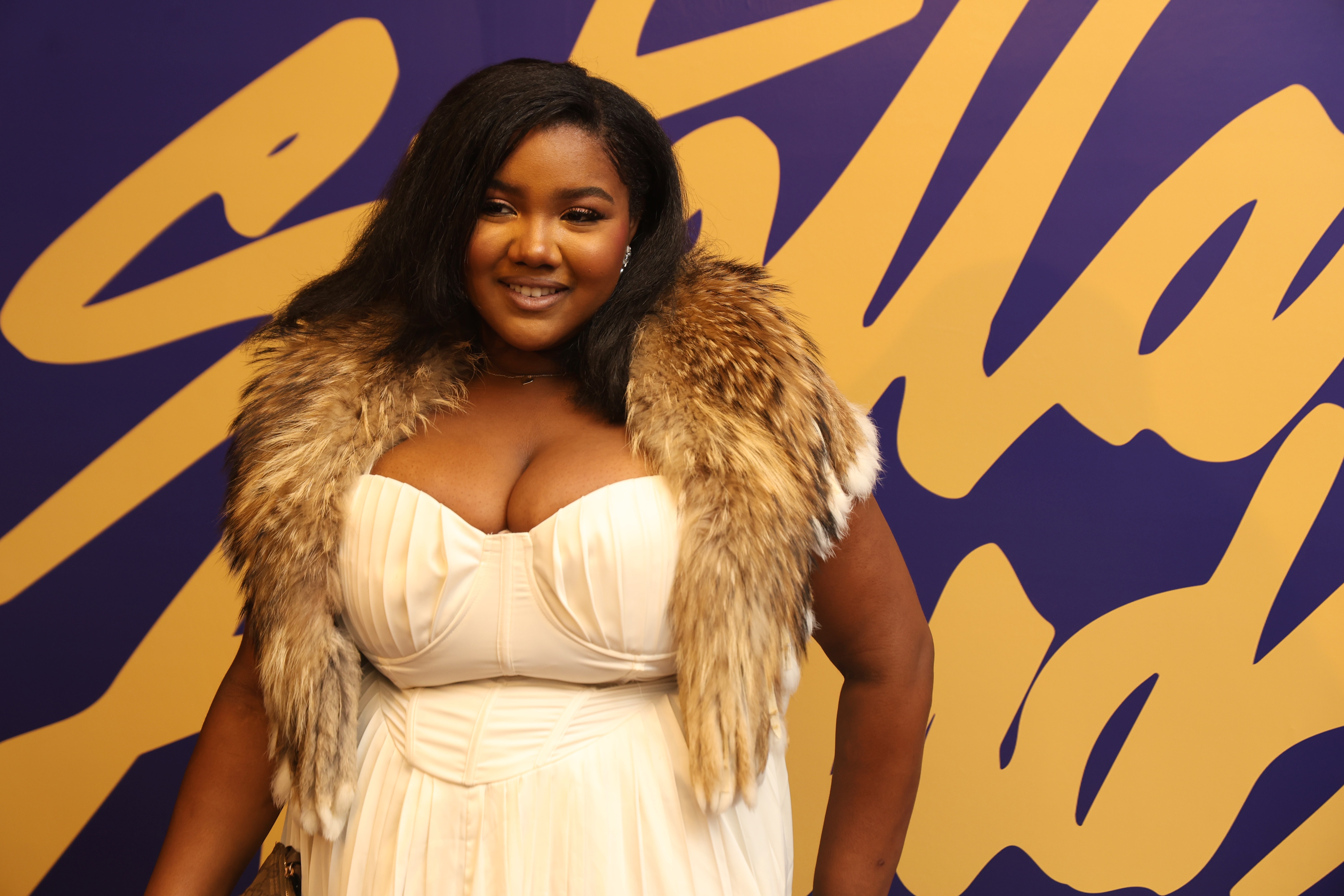 A smiling woman wearing a fur-trimmed white dress stands in front of a vibrant purple and yellow background.
