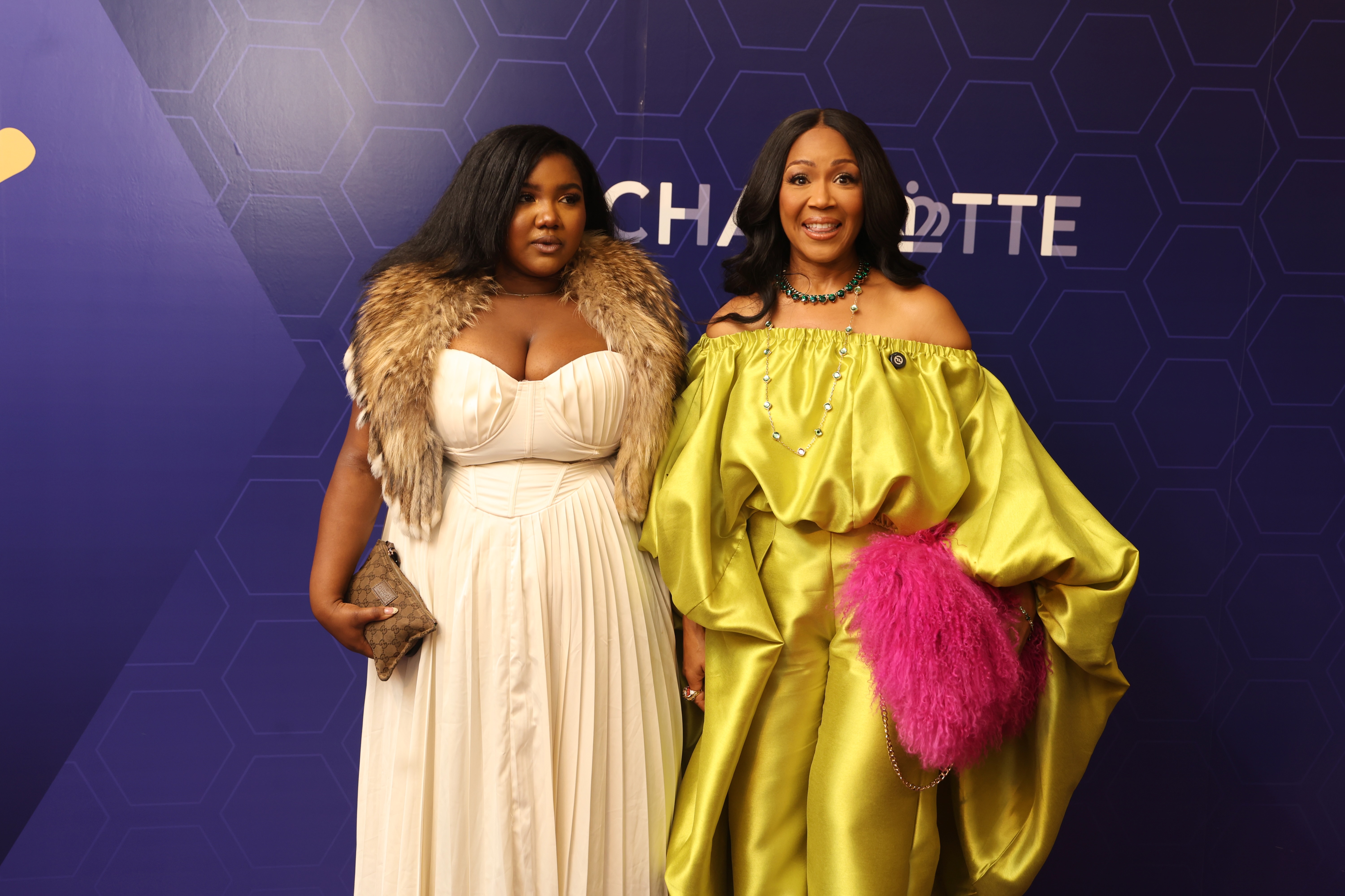 Two women wearing glamorous dresses posing in front of a purple and white backdrop with the "Charlotte" logo.