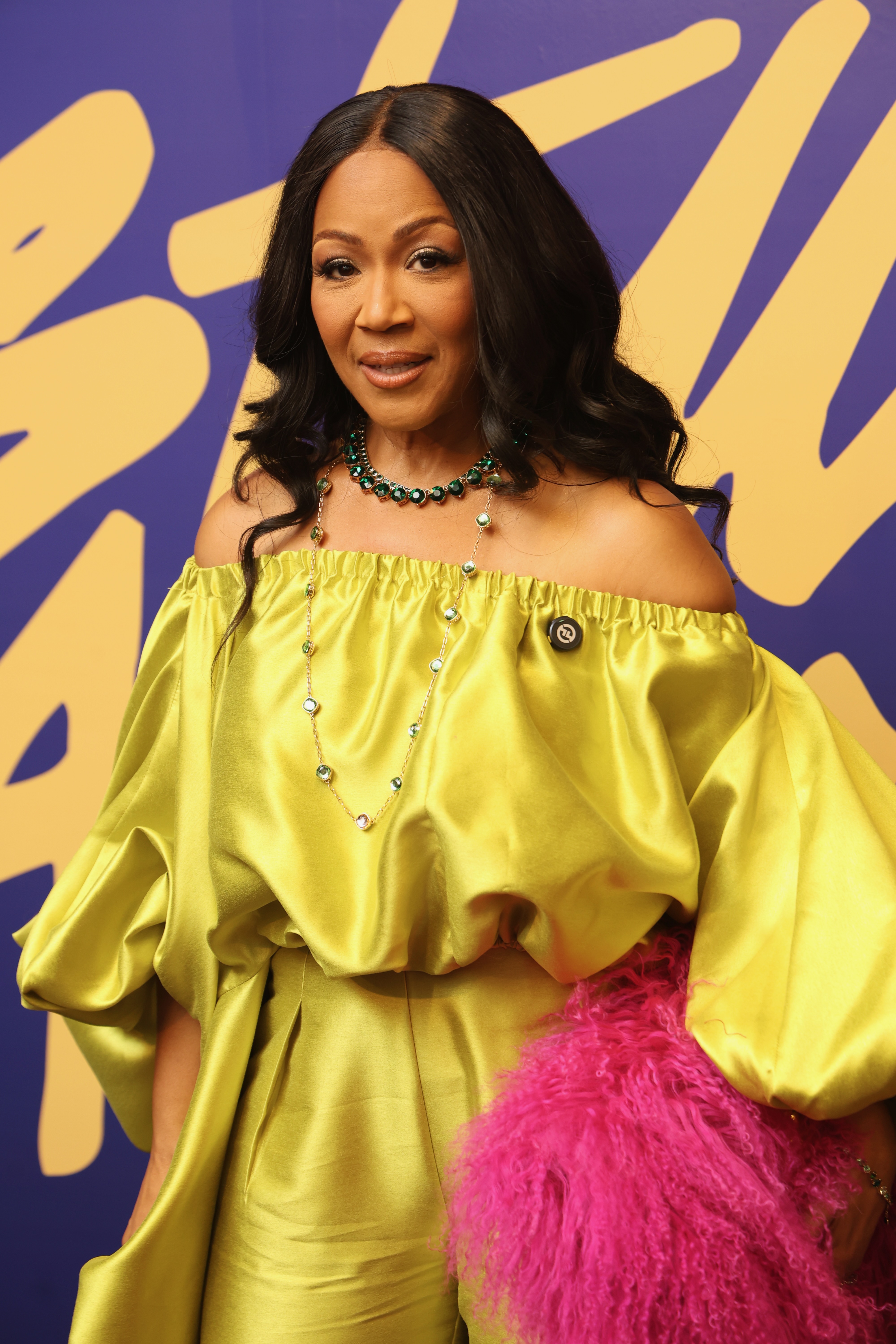 A woman wearing a bright yellow off-the-shoulder dress and a statement necklace stands in front of a purple and yellow abstract background.