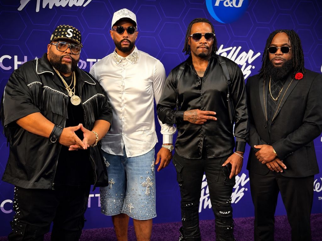 Four men in formal attire posing together, wearing sunglasses and jewelry, in front of a backdrop with "P&D" and "Killin' Them" branding.
