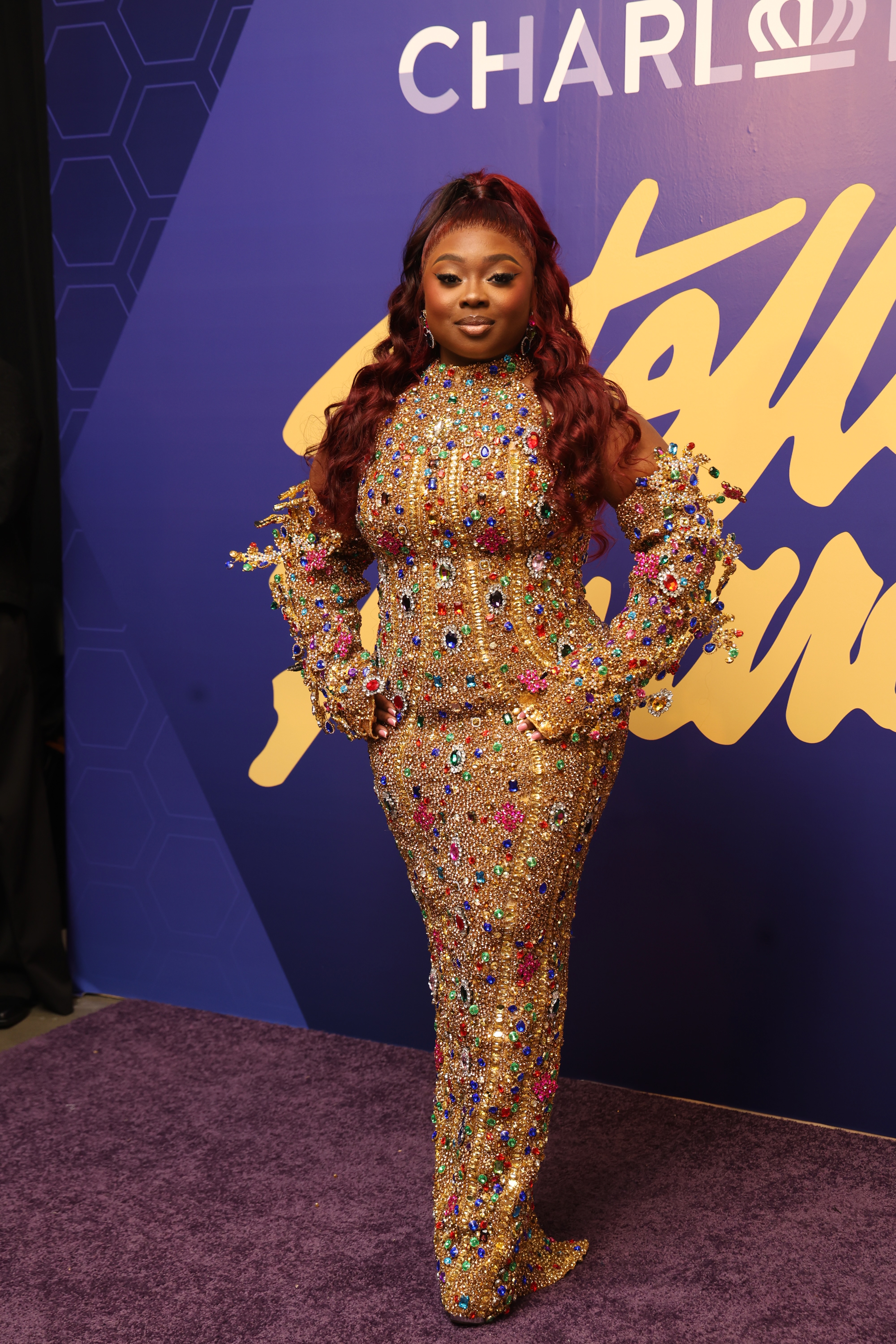 A person wearing a gold, bejeweled dress and standing in front of a purple backdrop with the text "CHARLI" displayed.