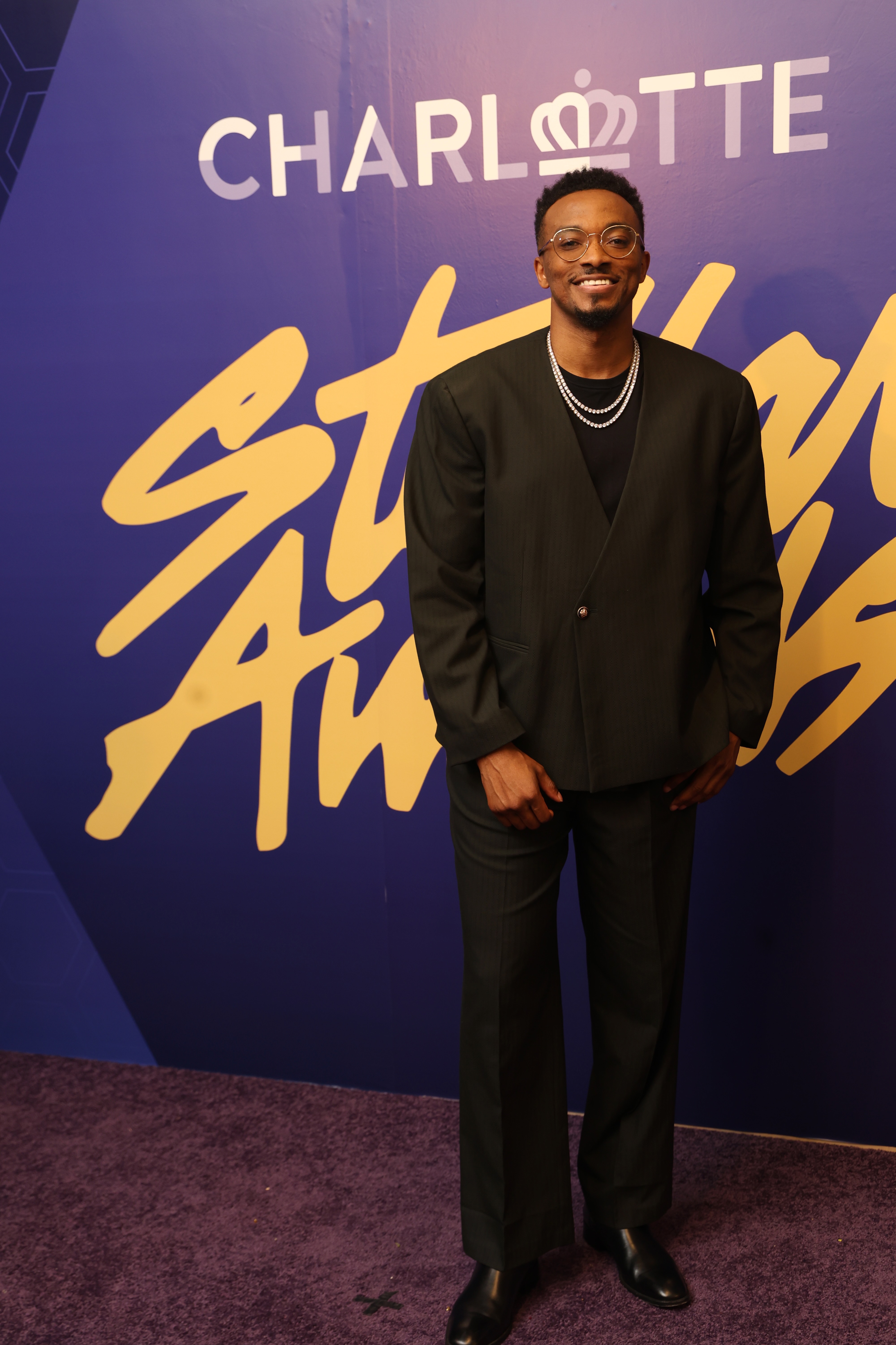 A smiling man wearing a black suit stands in front of a purple background with the text "CHARLOTTE" and "STORY AWARD" displayed.
