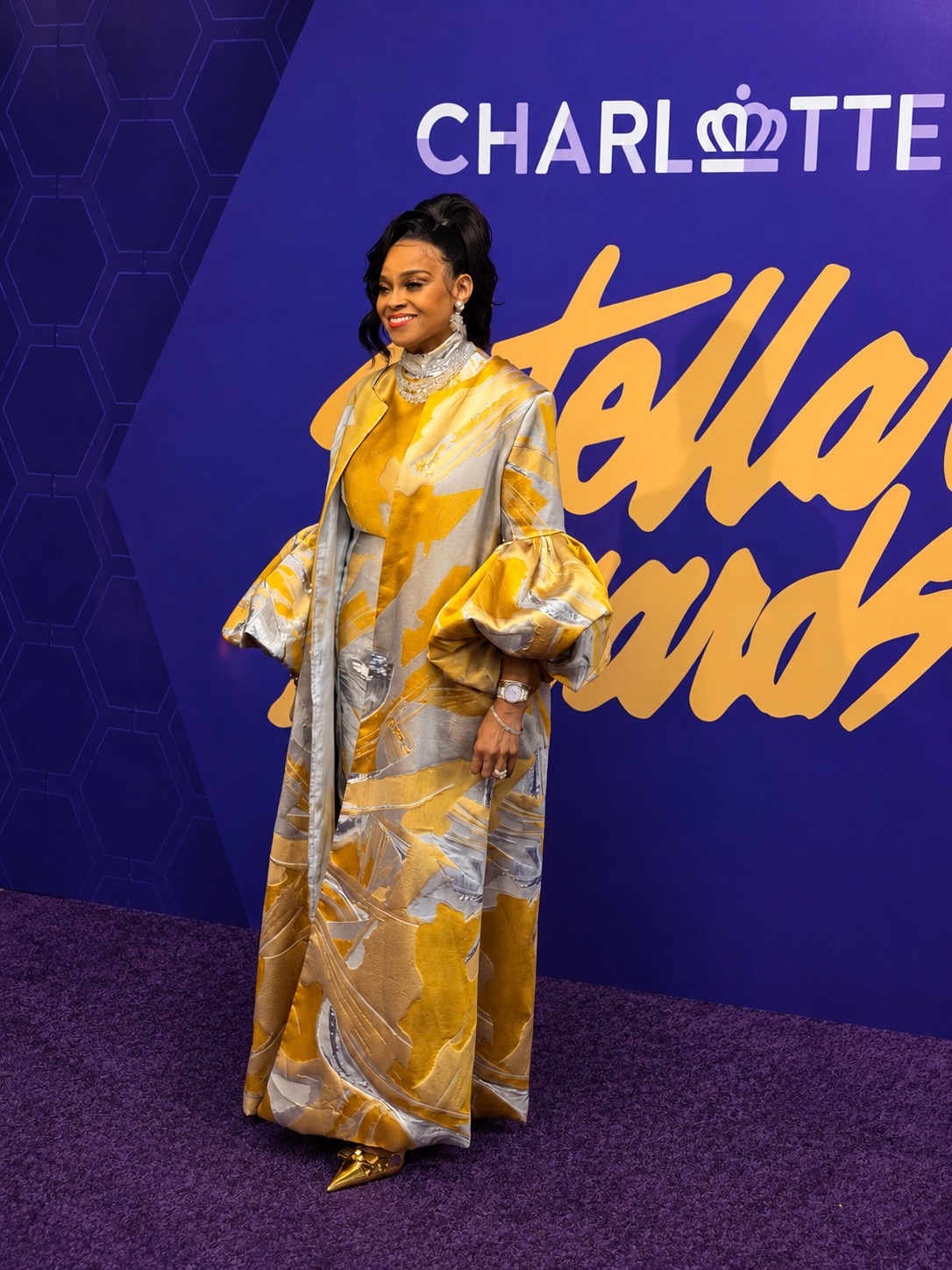 A woman in a vibrant yellow and gray patterned dress stands in front of a large "Charlotte" logo on a blue background, smiling at the camera.