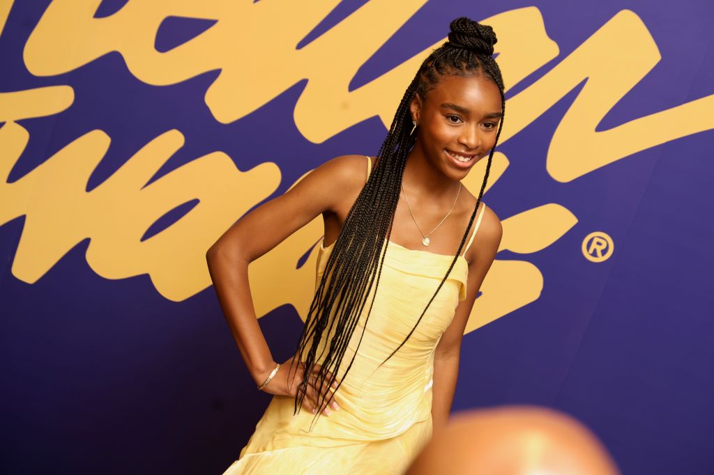 A smiling young woman with braided hair wearing a yellow dress stands in front of a purple and yellow abstract background.