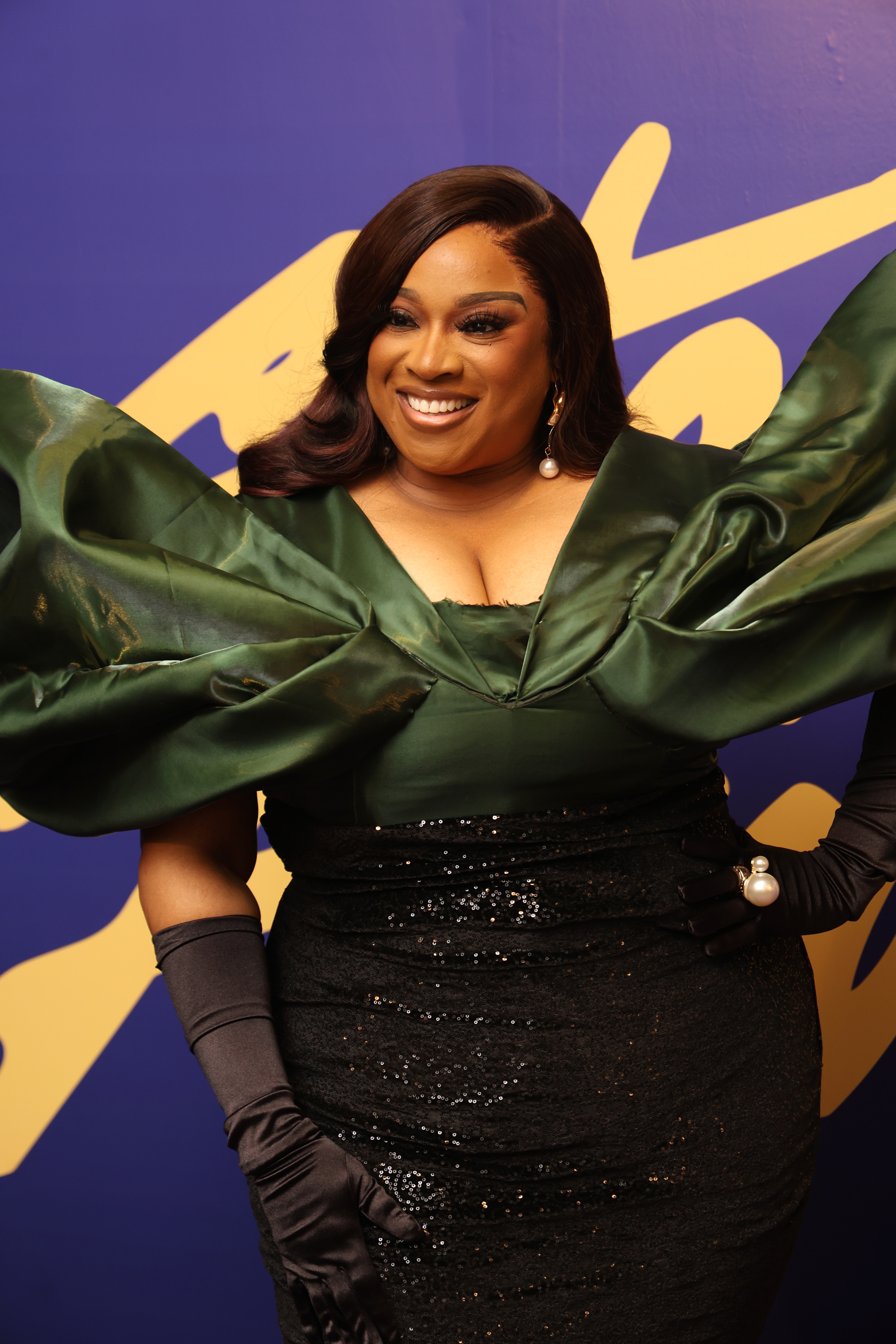 A smiling woman wearing a green dress with puffed sleeves stands against a purple and yellow background.