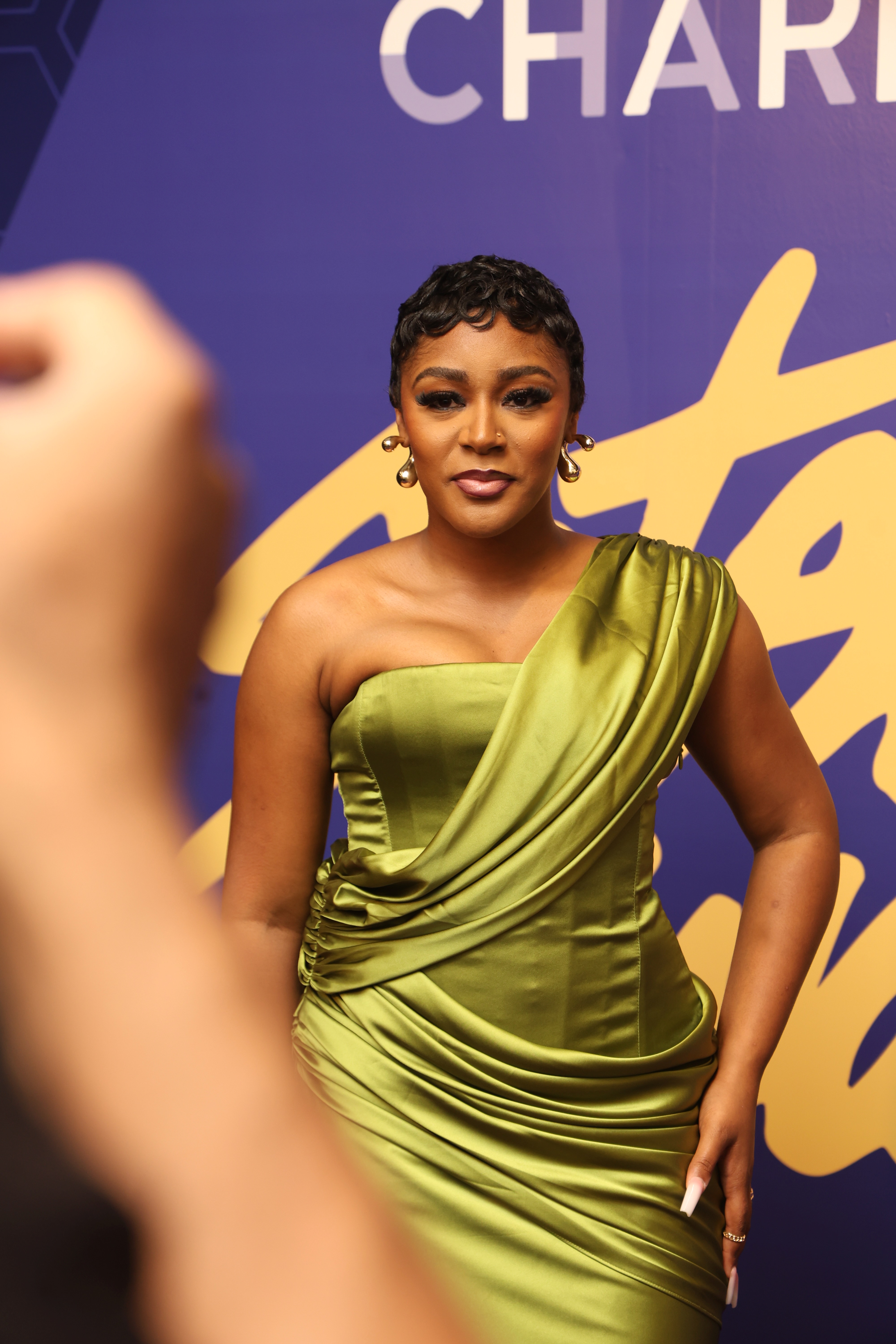 A woman in a green one-shoulder dress stands in front of a purple and yellow backdrop with the word "CHART" visible.