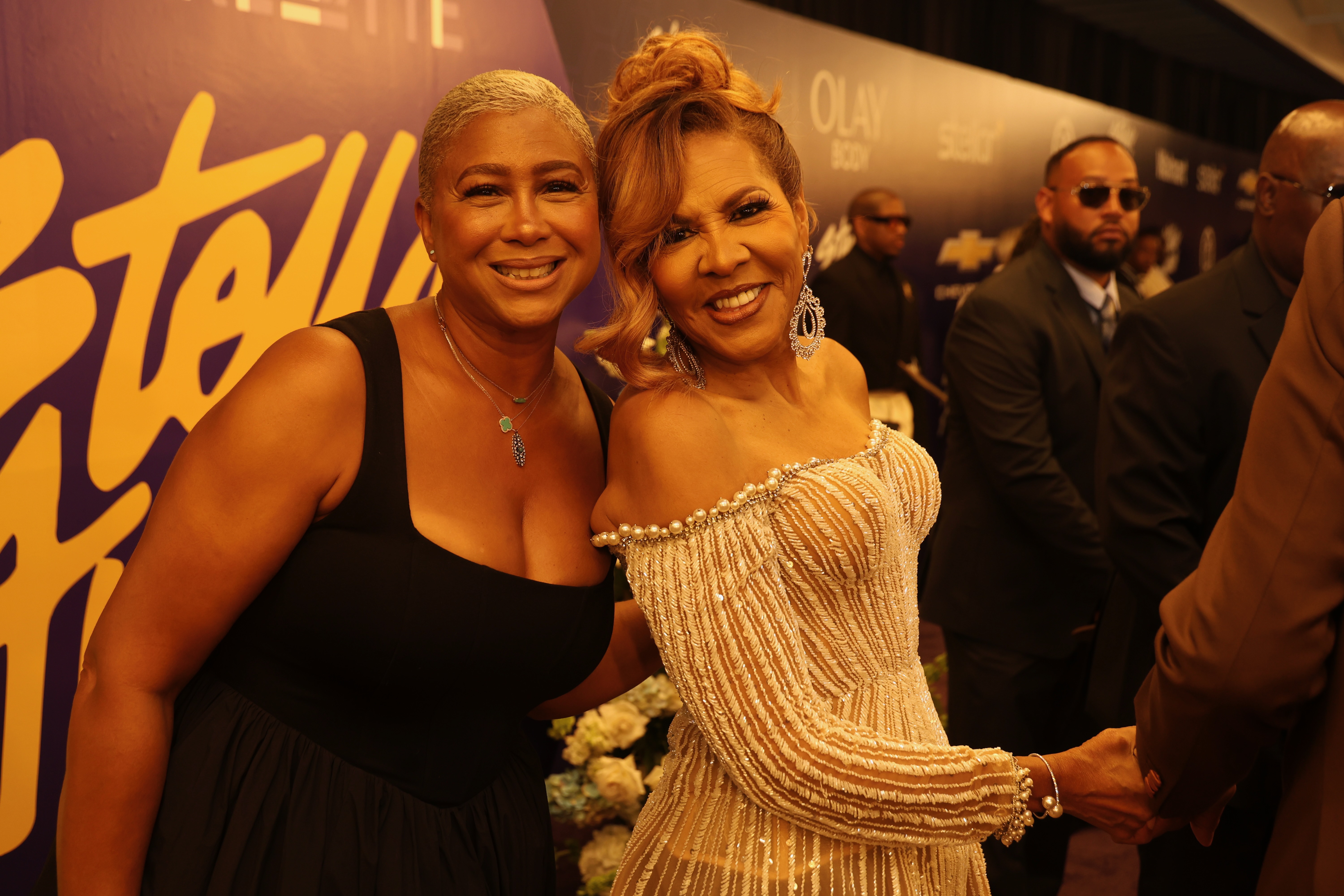 Two smiling women posing together at an event, one wearing a black dress and the other a gold sequined gown.