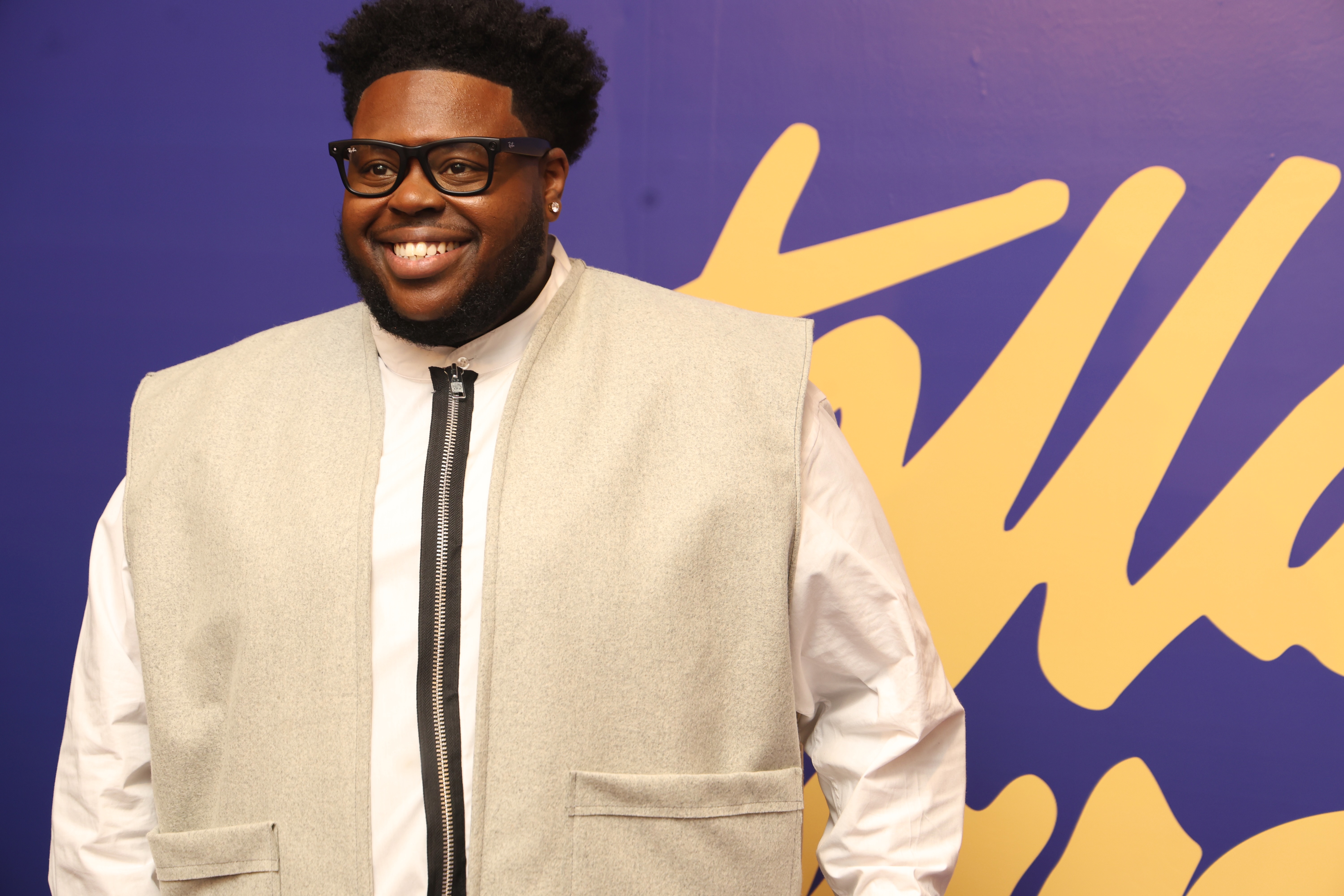 A smiling man wearing glasses and a white suit jacket stands in front of a purple and yellow abstract background.