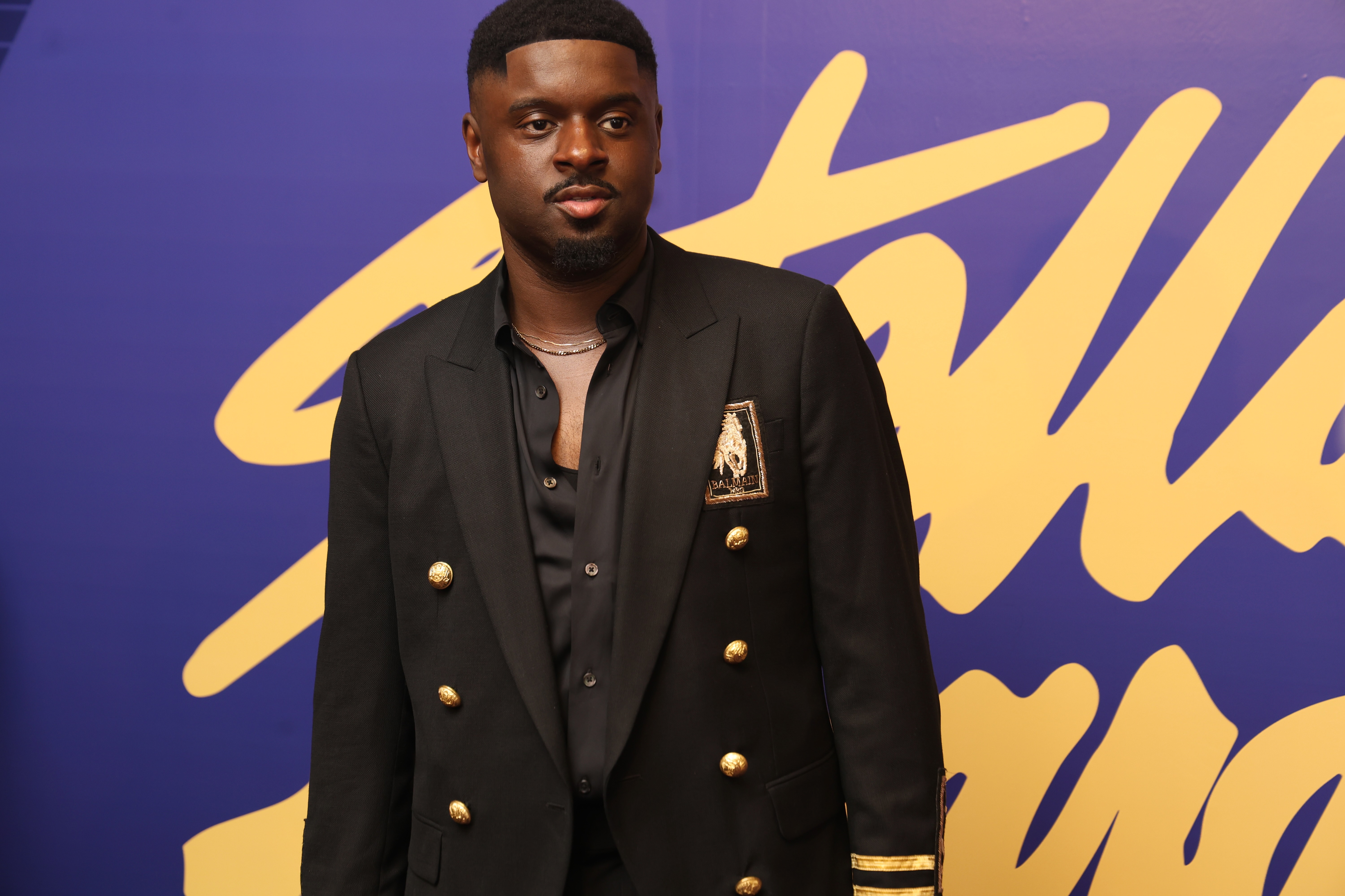 A man wearing a black suit jacket with gold buttons stands in front of a purple and yellow background.
