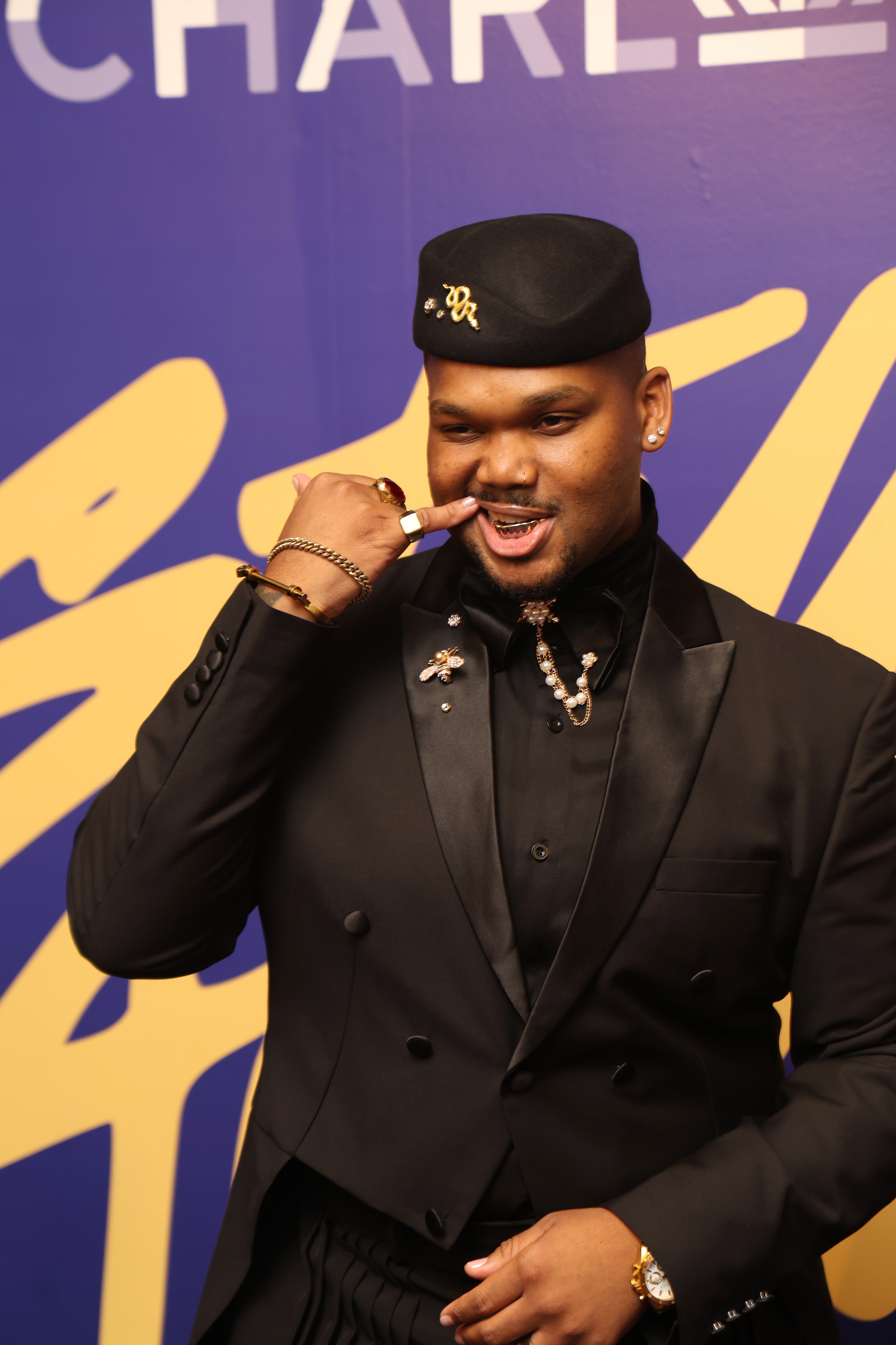 A man in a black suit and hat making a gesture with his hand in front of a purple and yellow background with the word "CHART" visible.