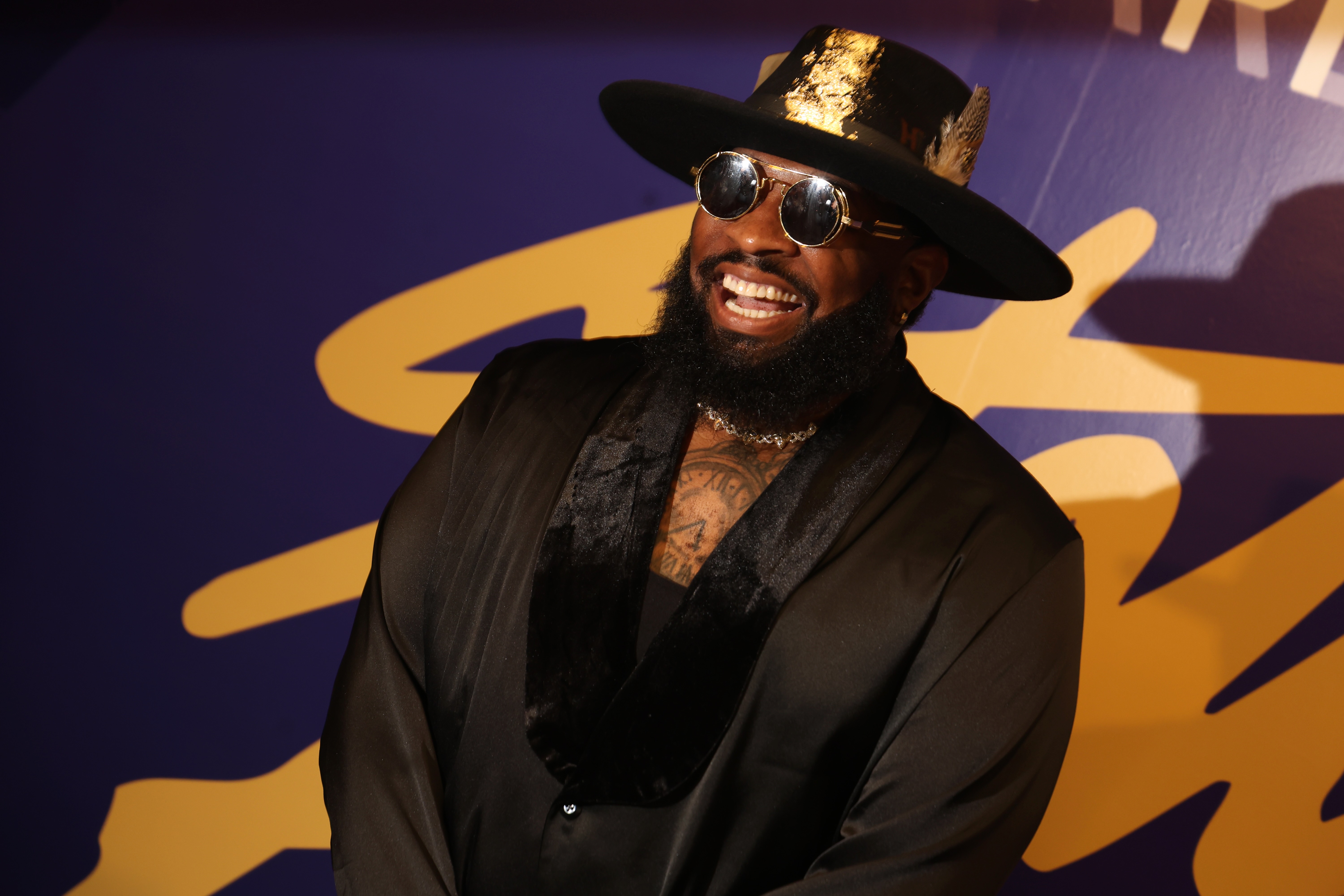 A smiling man with a beard and sunglasses wearing a black hat and coat stands in front of a colorful abstract background.
