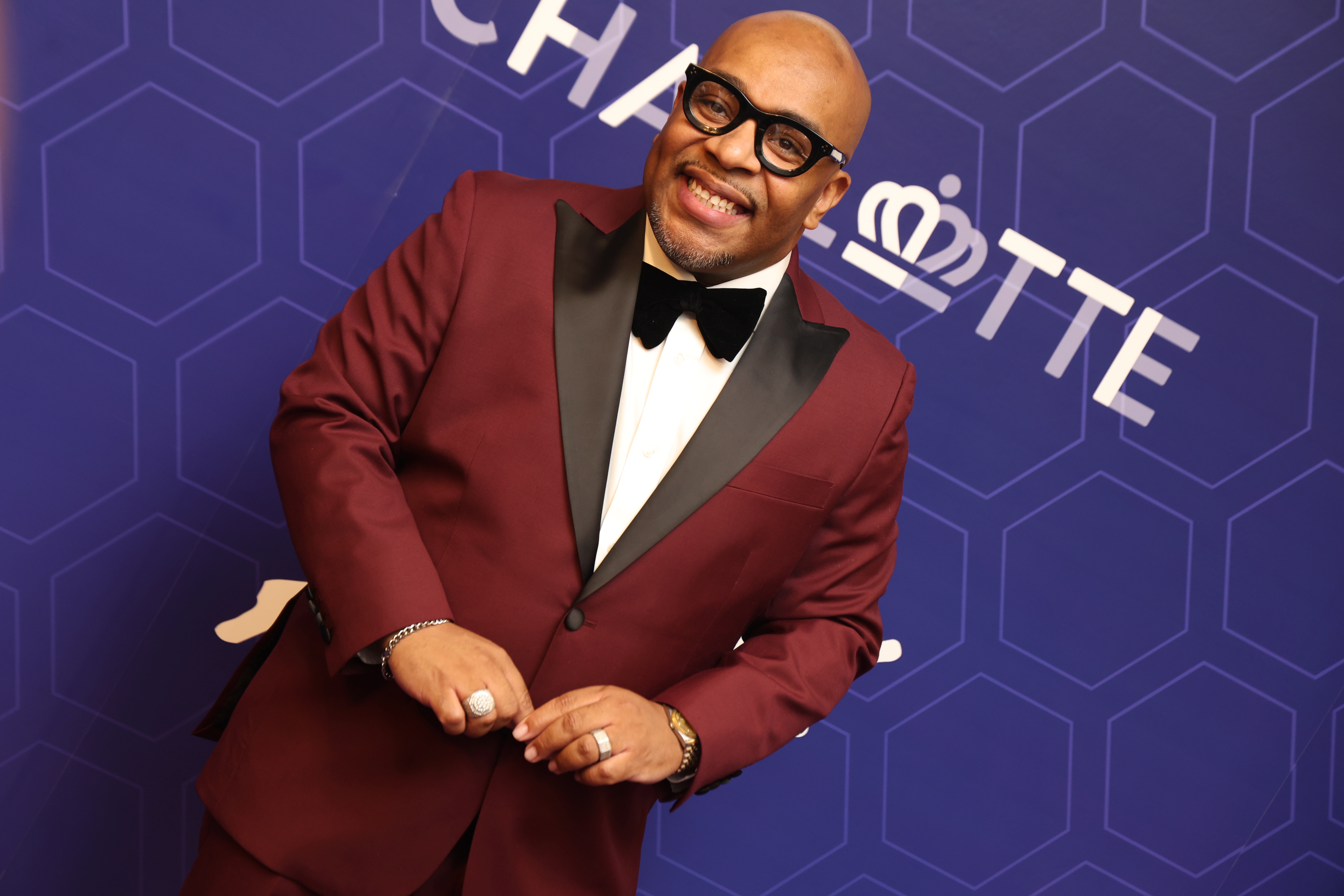 A smiling man wearing a red tuxedo jacket and bowtie stands in front of a purple backdrop with a "Charlotte" logo.